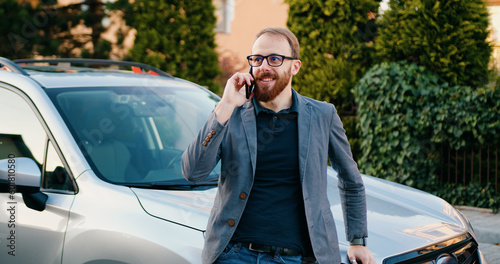 Stylish businessman talking on cell phone answering business call when leans on her modern car which is parked by area house. Outdoors
