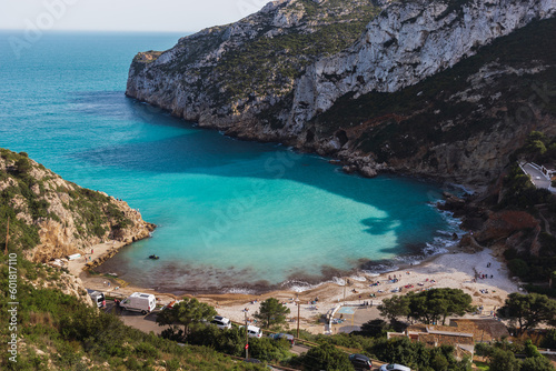 Views of the turquoise waters of Cala Granadella over the Mediterranean Sea. Costa Blanca, Javea, Alicante, Spain. On a sunny day with people on the beach and many cars around.