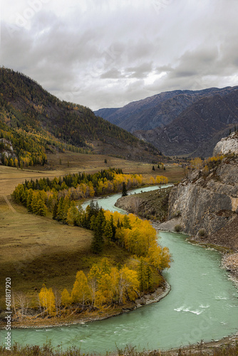 Vertical photo of panorama of an autumn landscape with yellow and green trees on the shore of a blue curving river flowing near cliffs and mountains in overcast weather with cloudy sky during the day