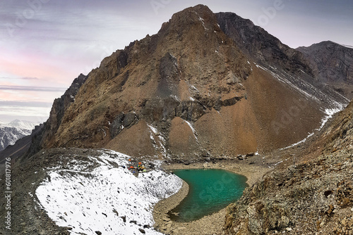 A green lake on top of a mountain with white snow and a climbing camp in the early morning with a cold sky