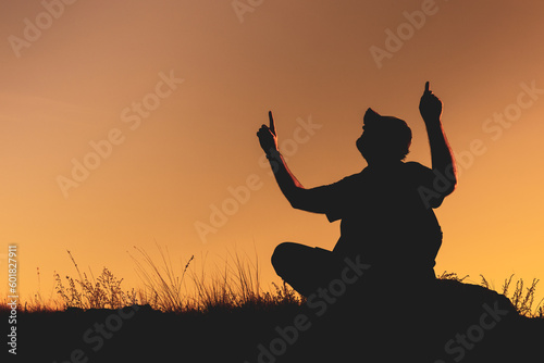 the silhouette of a man sitting outside on the grass and raising two hands up