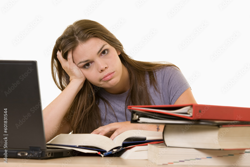 Young woman sitting in front of laptop beside a pile of thick textbooks while reading one with a frustrated stressed expression Young woman sitting in front of laptop beside a pile of thick textbooks while reading one with a frustrated stressed expression