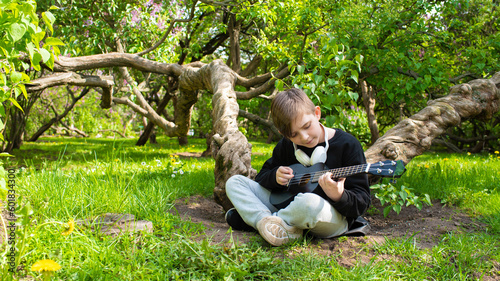 a little boy plays a small black ukulele, a guitar in a blooming garden, a park on a sunny day	
