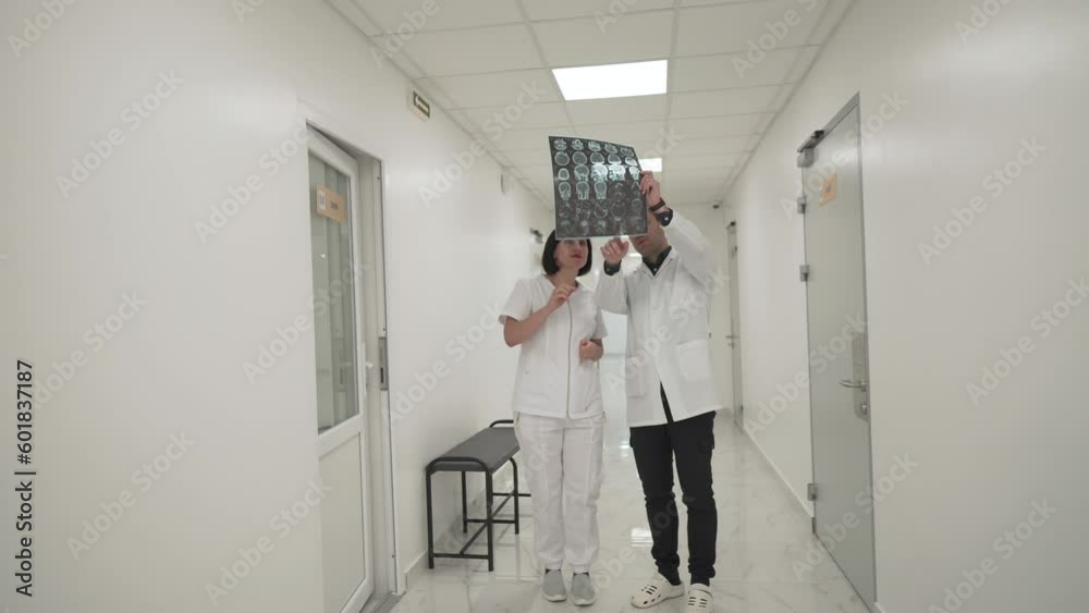 Teamwork in hospital. Two doctors move quickly across corridor of clinic and discuss an MRI scan of head of patient. Head doctor and assistant discuss CT scan of brain tumor patient in the hallway. Teamwork in hospital. Two doctors move quickly across corridor of clinic and discuss an MRI scan of head of patient. Head doctor and assistant discuss CT scan of brain tumor patient in the hallway.