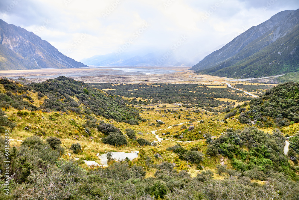 Blick durch ein neuseeländisches Tal gesäumt von Bergen mit grünen Landschaften bei regnerischem Wetter.