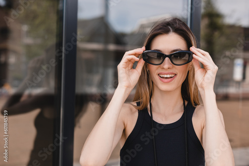 Pensive blonde woman in black t shirt put off black glasses and posing on office building background. Outdoor shot of happy business lady with teeth smile look happy.