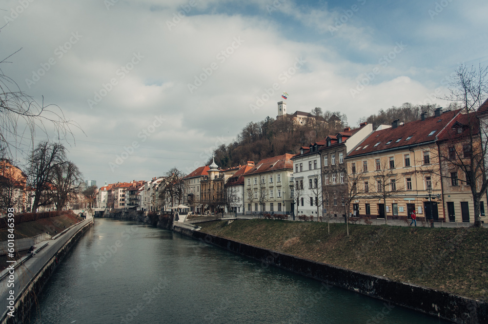 This photo captures the Ljubljana river, historical buildings, and castle during a cloudy day at the end of winter, showcasing the beauty and charm of the Slovenian capital.