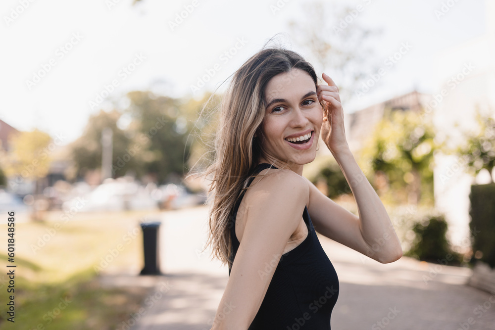 Beautiful woman smiling and look at camera in countryside. Outdoor portrait of a smiling blonde girl. Happy cheerful girl laughing at green city, look fashion and show toothy smile. She turn around.