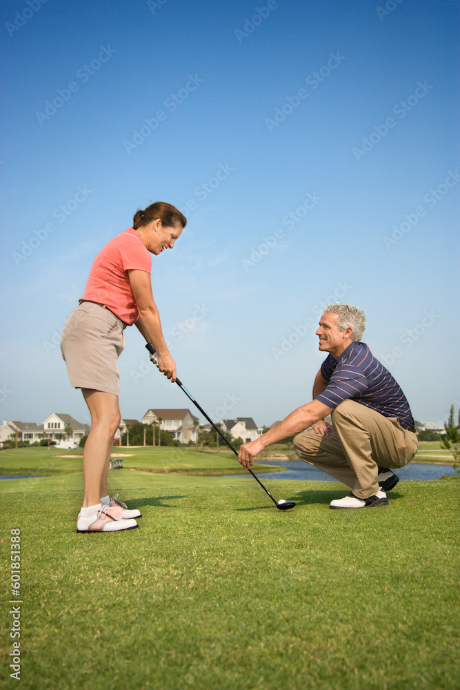 Caucasion mid-adult woman holding golf club while mid-adult man kneels holding club teaching.