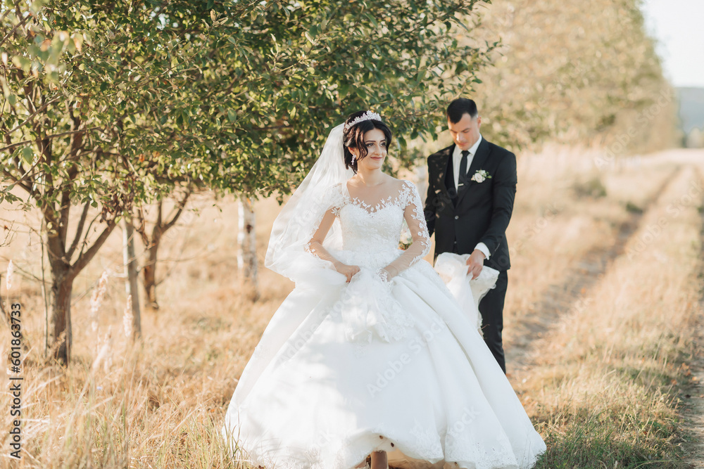 Wide-angle portrait of a bride and groom walking against a background of trees and a field. The bride is in front of the groom, the groom is holding the train of the dress behind her. 