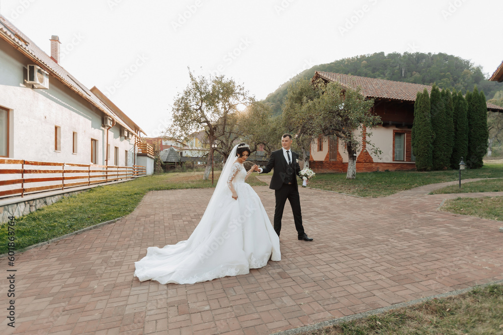Wedding photo. The groom in a black suit and the bride in a long white dress walk along the stone path holding hands, against the background of huts. Side view. Portrait. Beautiful crowns. 