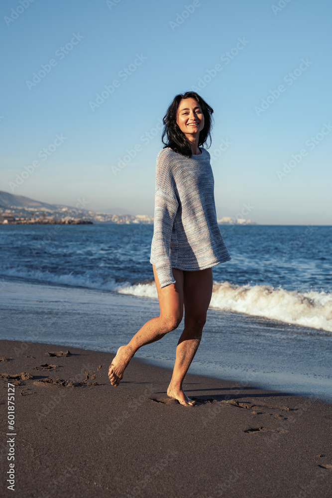 young beautiful brunette girl walks on the sand on the seashore in a swimsuit and a sweater