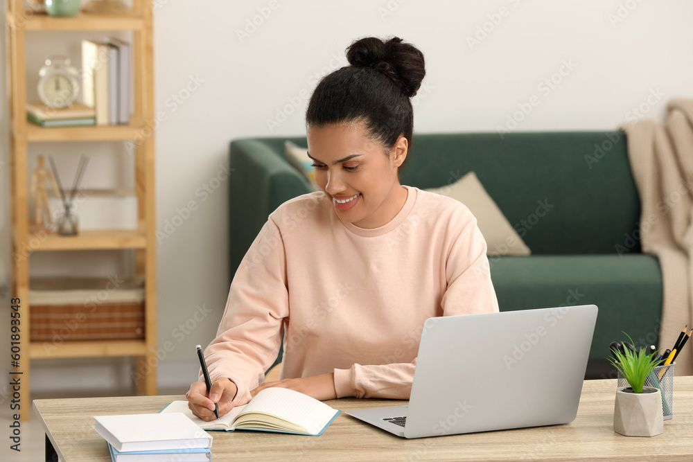 Beautiful African American woman writing in notepad near laptop at table in room Beautiful African American woman writing in notepad near laptop at table in room