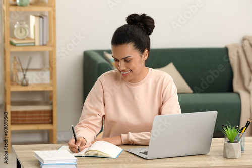Beautiful African American woman writing in notepad near laptop at table in room
