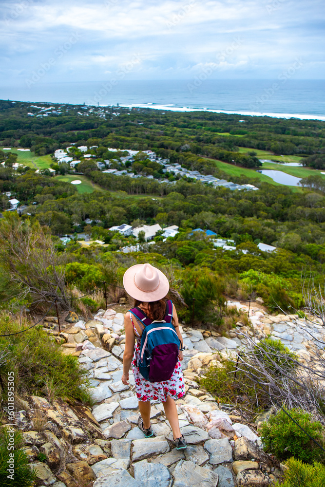 beautiful girl in a hat walking down the trail from mount coolum with panorama of sunshine coast and coolum beach in the background; hiking in queensland, australia