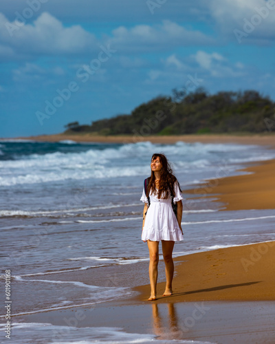 beautiful long haired girl in white dress walking lonely on the empty beach; walking on pristine beach with orange sand in deepwater national park near agnes water, australia