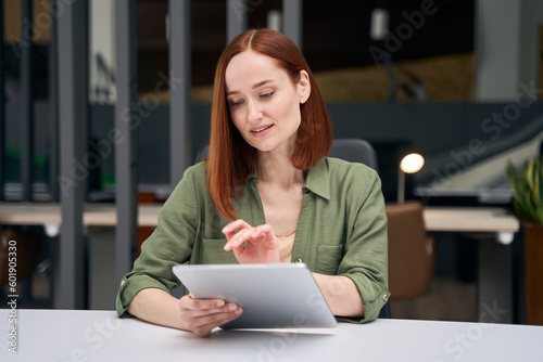 Confident smiling red haired businesswoman using digital tablet working online in modern office. Technology concept. Successful freelancer sitting at workplace