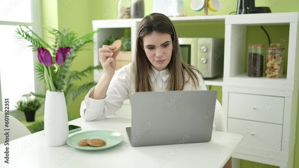 Young beautiful hispanic woman eating cookies using laptop at dinning room