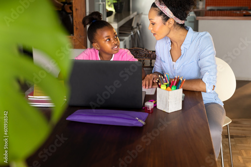 Smiling african american mother helping happy daughter doing schoolwork using laptop at home