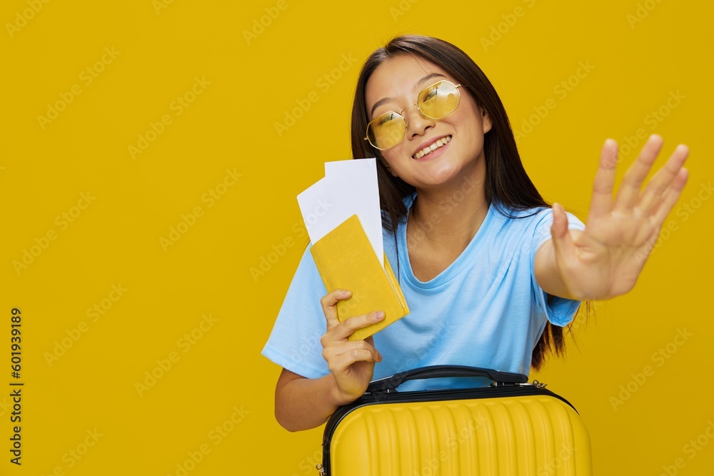 Asian woman traveling with yellow suitcase and tickets with passport in hand, tourist traveling by plane and train with luggage on yellow background in blue T-shirt and jeans Asian woman traveling with yellow suitcase and tickets with passport in hand, tourist traveling by plane and train with luggage on yellow background in blue T-shirt and jeans
