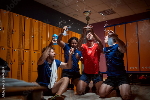 Group of basketball players holding up golden trophy and celebrating success in the dressing room, shouting and splashing water.
