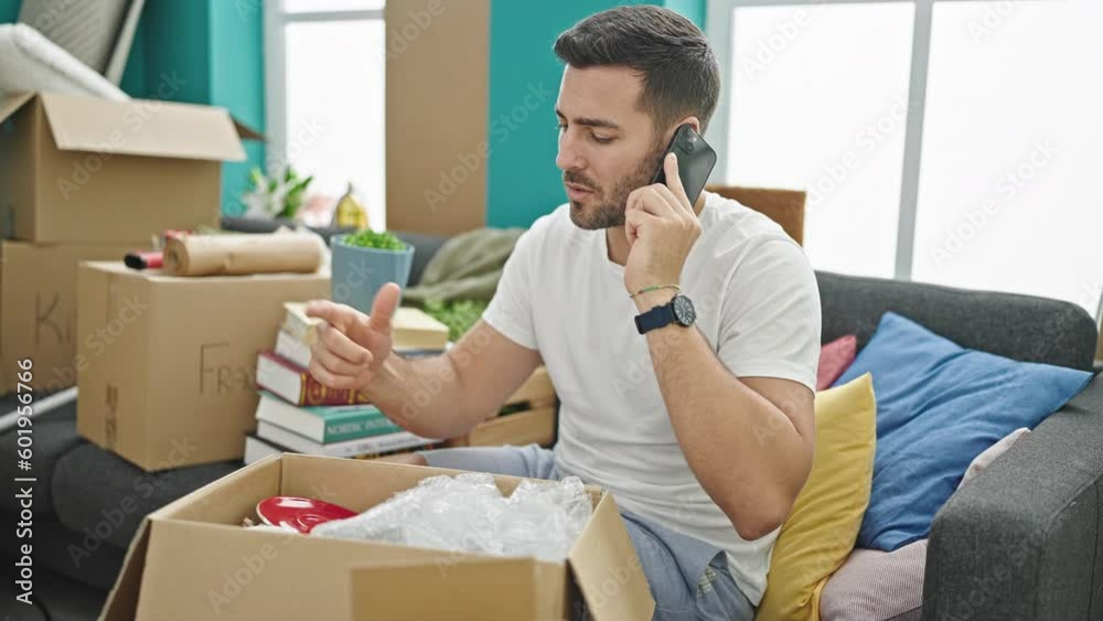 Young hispanic man unpacking cardboard box talking on smartphone at new home