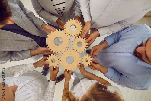 Group of creative business people joining together wooden gears at workplace in office. Top view of colleagues connecting gears of different sizes together as symbol of cooperation and teamwork.