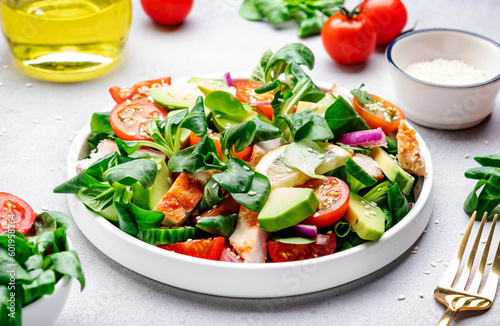 Chicken and vegetables salad with tomatoes,  avocado, cucumber, onion, lamb lettuce and sesame seeds on gray table background, top view