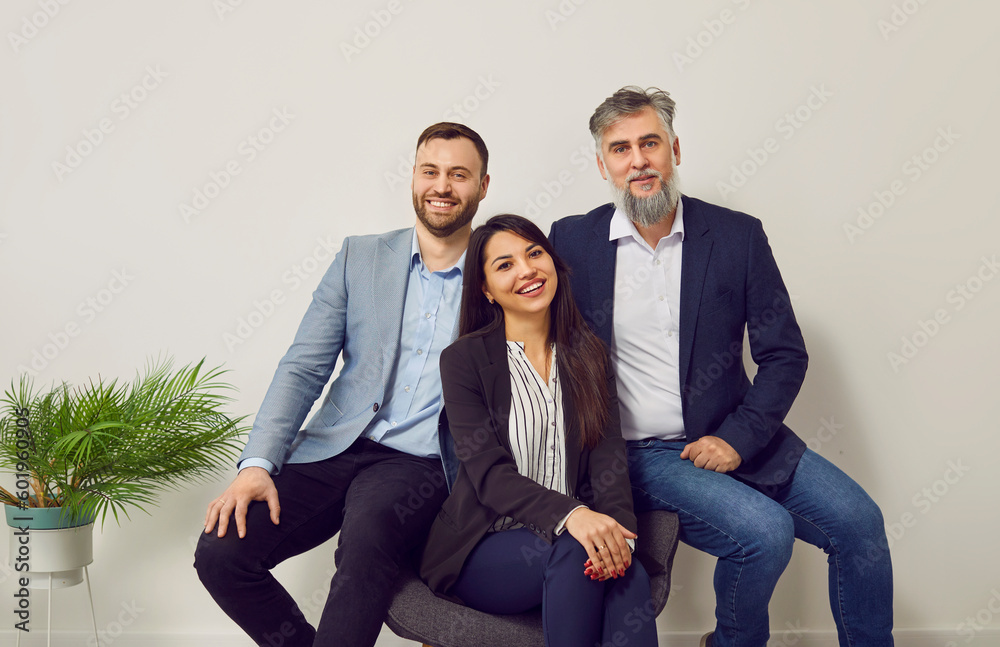Group of smiling business workers posing together in office. Company staff members, employees in formal wear sitting on one chair together against gray wall face looking at camera Group of smiling business workers posing together in office. Company staff members, employees in formal wear sitting on one chair together against gray wall face looking at camera