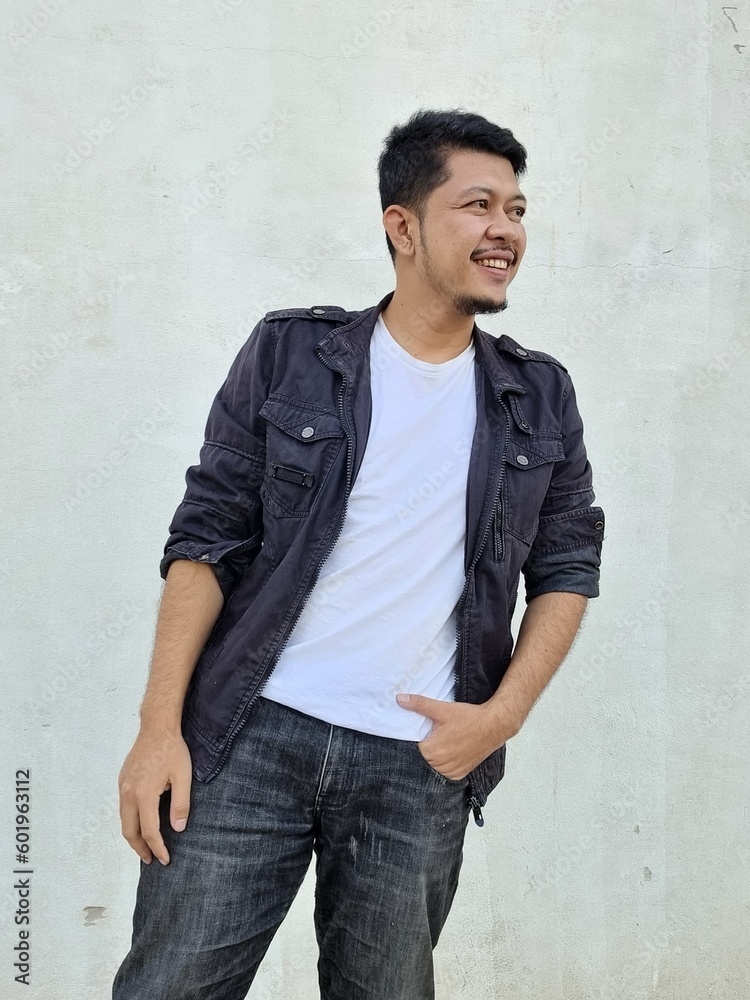 Cool modern young hipster man with a stylish hairstyle in a white T-shirt puts on a vintage black denim jacket. Attractive urban guy posing near a white wall. Fashionable spring menswear. Cool modern young hipster man with a stylish hairstyle in a white T-shirt puts on a vintage black denim jacket. Attractive urban guy posing near a white wall. Fashionable spring menswear.