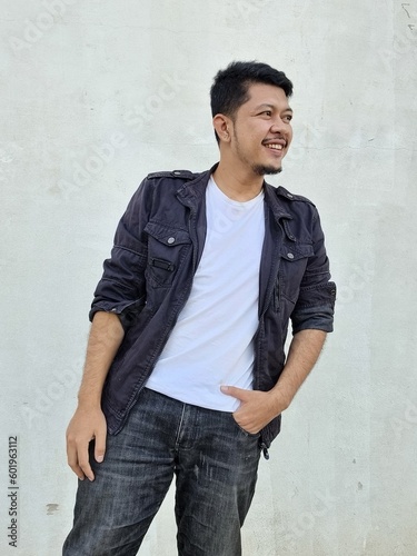 Cool modern young hipster man with a stylish hairstyle in a white T-shirt puts on a vintage black denim jacket. Attractive urban guy posing near a white wall. Fashionable spring menswear.
