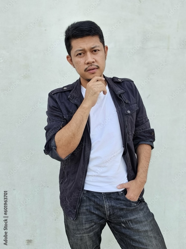 Cool modern young hipster man with a stylish hairstyle in a white T-shirt puts on a vintage black denim jacket. Attractive urban guy posing near a white wall. Fashionable spring menswear. Cool modern young hipster man with a stylish hairstyle in a white T-shirt puts on a vintage black denim jacket. Attractive urban guy posing near a white wall. Fashionable spring menswear.