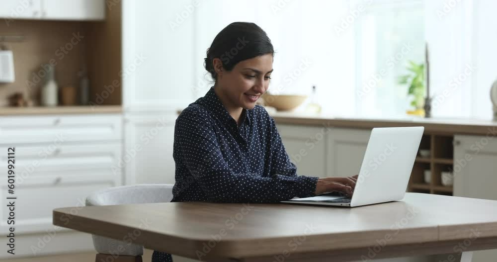 Millennial Indian woman sitting in kitchen and working on laptop, smile while typing message on notebook keyboard, share message, doing freelance telecommute work from home using internet connection