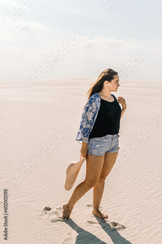 a girl in a black T-shirt and denim shorts in a white cape with a blue pattern, a cardigan, a poncho with a hat on her head in the desert, on the beach on the sand