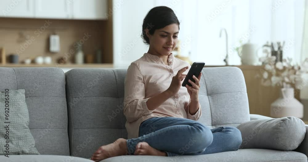Carefree Indian woman sit on sofa in living room with smartphone, check email, chat in social media, share message, web surfing, search information spend leisure at home, make purchase use mobile app Carefree Indian woman sit on sofa in living room with smartphone, check email, chat in social media, share message, web surfing, search information spend leisure at home, make purchase use mobile app