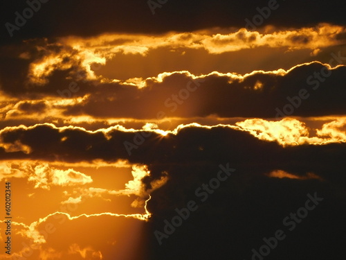 Angelic mesmerizing skyline at sunset with rows of grey large heavy clouds illuminated by the sun behind casting bright orange and white light around and between the clouds 
