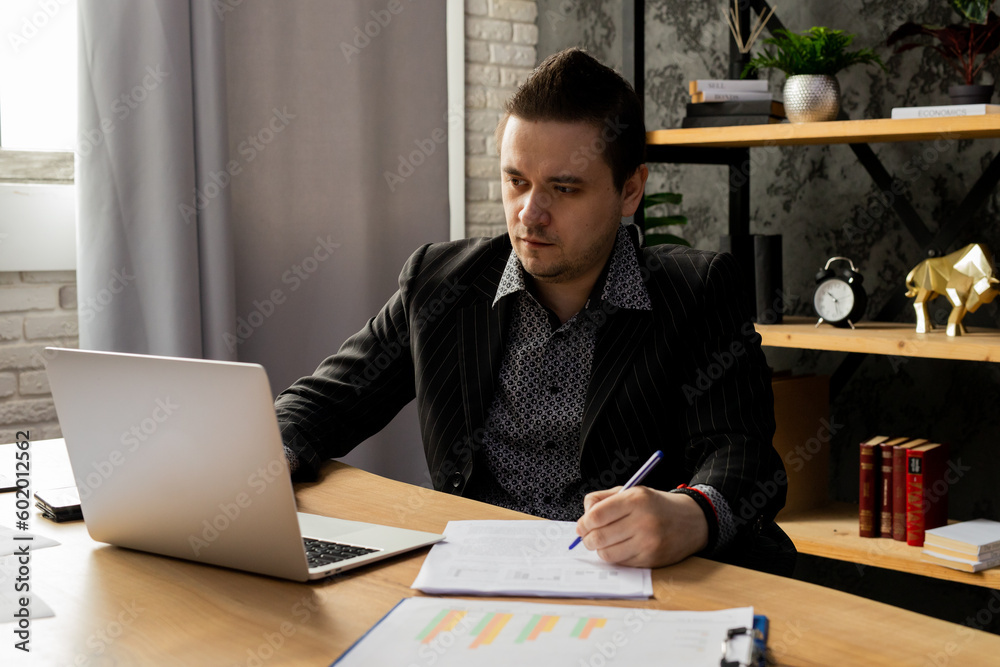 Fototapeta premium A young man in a business suit is working on a laptop while sitting in the office. A man in a black jacket sits in front of a laptop and fills out paperwork.