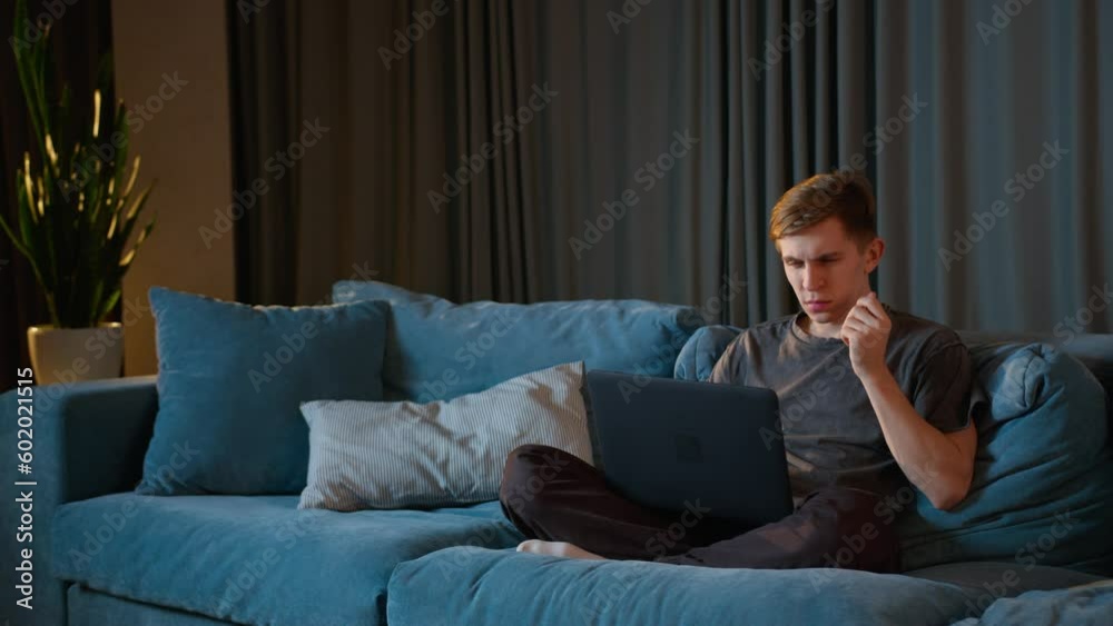 a serious young man working on his laptop while sitting on the couch. Handsome young man in home clothes working at his computer from home. a serious young man working on his laptop while sitting on the couch. Handsome young man in home clothes working at his computer from home.