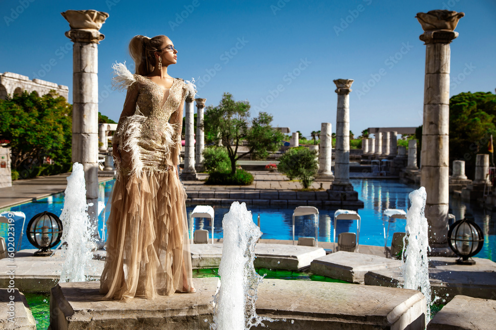 Glamour, stylish elegant woman in long gown feather dress is posing need the pool in luxury hotel. Female model in amazing long dress. Outdoor shoot. Vogue. Couture.