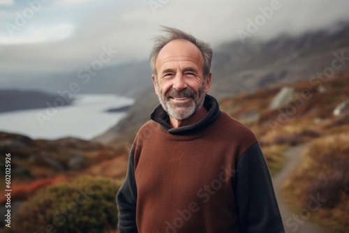 Portrait of a handsome senior man with gray hair, wearing a brown sweater, standing on a mountain trail and looking at the camera.