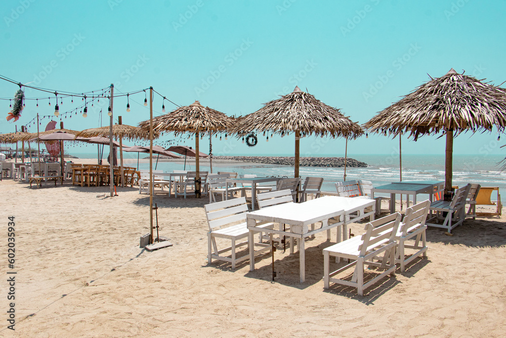 Wooden seaside parasols (Straw Umbrellas) and seats with table on the sand beach landscape in clear blue sky for vacation concept in summer holiday