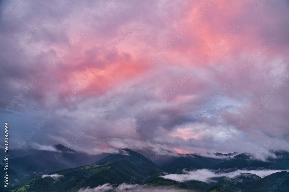 Mountains in low clouds at sunset in summer. view of mountain hills with green trees in fog and colorful sky with red clouds at dusk. Beautiful landscape.