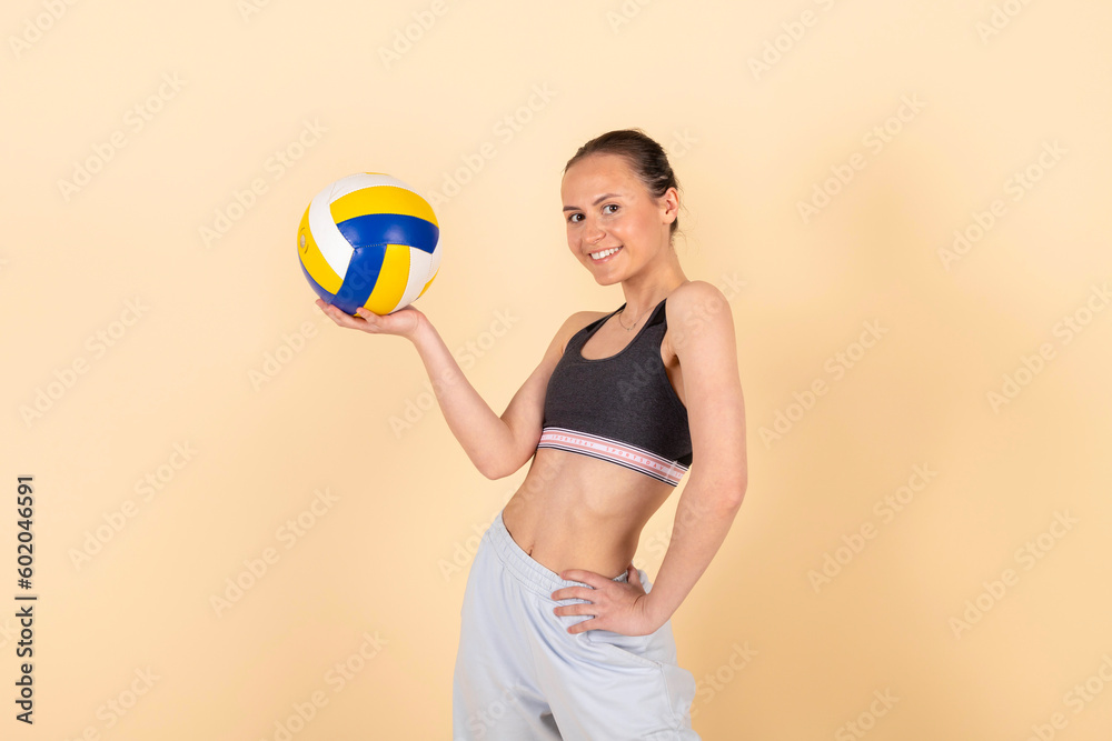 Cute slim brunette girl in sportswear posing with a volleyball. Young athlete with a ball. Isolated on pastel orange, peach background.