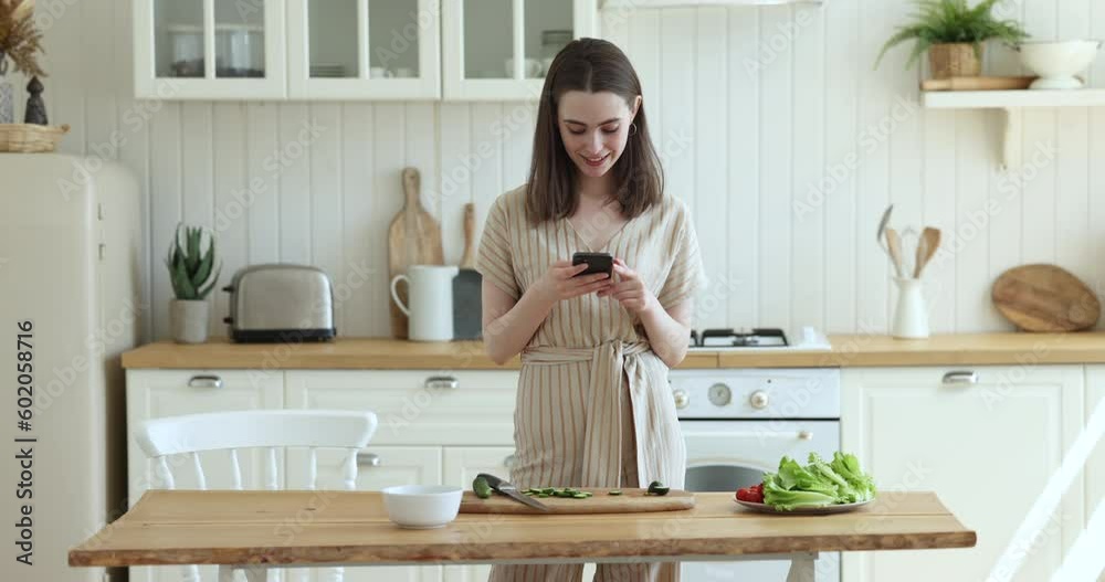 Happy young adult girl using smartphone in kitchen, typing, consulting cook blog, reading online recipe, preparing organic healthy salad for dinner, cutting fresh vegetables, taking picture of table Happy young adult girl using smartphone in kitchen, typing, consulting cook blog, reading online recipe, preparing organic healthy salad for dinner, cutting fresh vegetables, taking picture of table