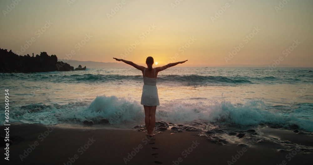 Woman silhouette raises hands up on black sand beach at sunset. Twilight orange sky. Adult girl relax watching powerful sea waves landscape. Woman silhouette raises hands up on black sand beach at sunset. Twilight orange sky. Adult girl relax watching powerful sea waves landscape.