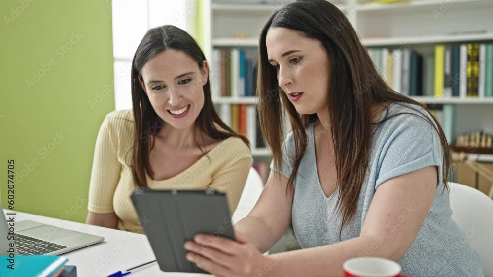 Two women sitting on table studying using laptop and touchpad at library university Two women sitting on table studying using laptop and touchpad at library university