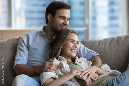 Cheerful positive young dating couple hugging with love, relaxing at home, resting on couch, looking away, thinking, dreaming, planning, laughing, smiling, enjoying leisure, healthy relationship