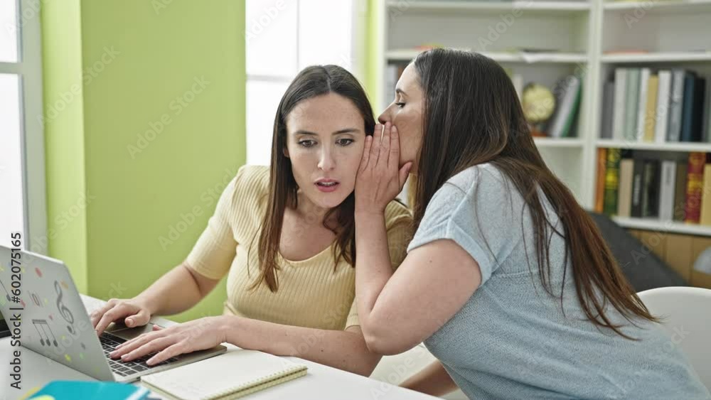 Two women sitting on table using laptop telling secret at library university Two women sitting on table using laptop telling secret at library university