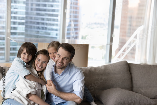 Happy attractive young parents piggybacking two cheerful cute kids, playing active games with children on sofa, laughing, smiling, looking at camera away. Candid family portrait with copy space