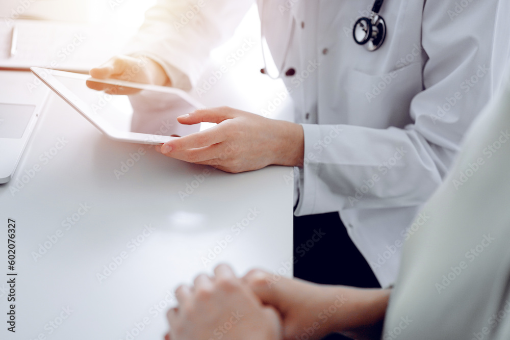 Doctor and patient sitting at the desk in clinic office. The focus is on female physician's hands using tablet computer, close up. Perfect medical service and medicine concept Doctor and patient sitting at the desk in clinic office. The focus is on female physician's hands using tablet computer, close up. Perfect medical service and medicine concept